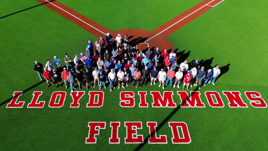 Former Trojan baseball players and Coach Lloyd Simmons gather on the field and pose for a group photo after the turf addition labeling Lloyd Simmons Field was revealed.